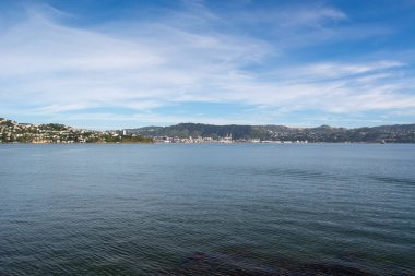 Sakin bir körfez manzarası, uzak tepeler ve açık mavi gökyüzünün altında bir şehir silueti. Su gökyüzünü yansıtır, barışçıl bir atmosfer yaratır..