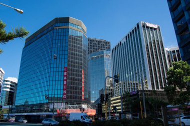 Yansıtıcı cam cepheli birkaç yüksek katlı ofis binasının yer aldığı modern bir şehir manzarası. Sahne, önplanda açık mavi bir gökyüzü ve yeşil bir şehir atmosferi yaratıyor..