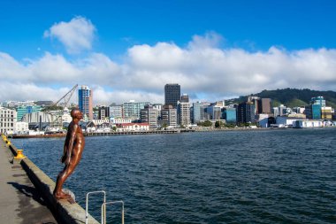 Canlı bir şehrin gökyüzüne bakan, modern binalar ve açık mavi gökyüzü olan bir adamın bronz heykeli. Su şehri yansıtıyor, sakin bir atmosfer yaratıyor..