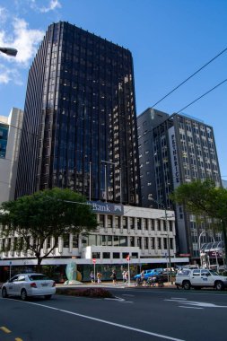 Modern ve klasik mimari içeren bir şehir manzarası. Ön planda beyaz yüzlü bir banka binası gösterilirken, arka planda uzun, karanlık gökdelenler yer alıyor. Sokaklarda yayalar ve araçlar görülüyor, bu da kentsel yaşamı gösteriyor..