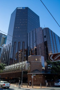Tepesinde ANZ banka logosunun bulunduğu yansıtıcı cam pencereli modern bir gökdelen. Bina kıtalararası otelin bitişiğinde, arka planda açık mavi bir gökyüzü var..