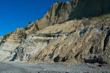 Açık mavi gökyüzüne karşı katmanlı tortu oluşumlarıyla kayalık yamaçlarda.