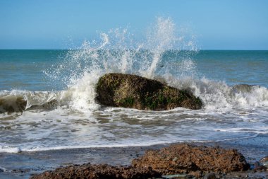 Büyük bir kaya kısmen okyanusa battı, dalgalar ona çarpıp bir sıçrama yarattı. Sahne, açık mavi gökyüzü ve nazik dalgalarla doğanın güzelliğini yakalar..