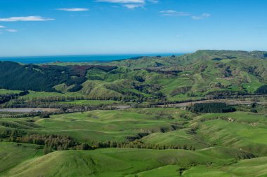 Berrak mavi gökyüzünün altındaki yemyeşil tepelerin ve vadilerin panoramik manzarası. Okyanus da uzaktan görülebiliyor. Manzara yuvarlanan bir araziye ve yeşilliklerin arasında kıvrılan bir nehre sahiptir..