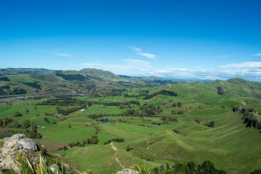 Berrak mavi gökyüzünün altındaki yemyeşil tepelerin ve vadilerin panoramik görüntüsü. Manzara, kırsal manzaranın güzelliğini gözler önüne seren yuvarlanan tepeler, ağaçlar ve dolambaçlı bir nehirden oluşuyor..