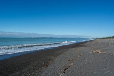 Siyah bir kumsal sahili sahnesi, kıyıda hafif dalgalar ve açık mavi bir gökyüzü. Ufuk parlak güneşin altındaki uzak kara parçalarını gösteriyor..
