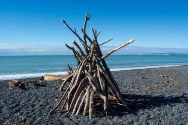 Okyanus ve arka planında açık mavi bir gökyüzü olan, siyah bir çakıl taşı plajında kırsal bir kaygan ağaç yapısı duruyor. Sahne bir huzur ve doğal güzelliği çağrıştırıyor..