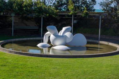 Çimen ve ağaçlarla çevrili dairesel bir gölette modern bir soyut heykel. Heykel, suya yansıyan, bembeyaz organik şekiller içeriyor..