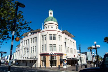 Yeşil kubbesi ve saat kulesi olan tarihi bir bina. Sanat deco mimarisi tarzında. Yapı, palmiye ağaçları ve sokak lambalarıyla çevrili açık mavi bir gökyüzüne karşı kurulmuştur..