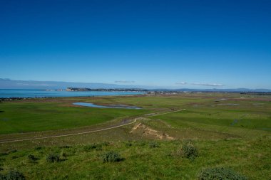 Bereketli yeşil tarlalar, sakin mavi bir deniz ve berrak mavi gökyüzünün altındaki uzak bir şehrin panoramik manzarası..