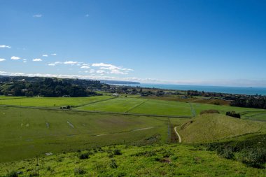 Bereketli yeşil tarlaların manzarası ve açık mavi gökyüzünün altındaki kıyı manzarası. Ufuk sakin bir denize sahiptir, uzaktaki küçük bir kasaba görülebilir..