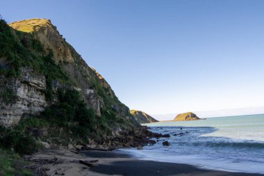 Engebeli kayalıklardan ve sakin bir denizden oluşan sakin bir kıyı manzarası. Kumsal dağınık kayalarla kumlu ve gökyüzü bulutsuz, barışçıl bir atmosfer yaratıyor..