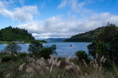 Parçalı bulutlu bir gökyüzünün altında yemyeşil tepelerle çevrili sakin bir göl. Suyun üzerinde küçük bir tekne görünür, ön planda uzun otlar doğal güzelliği artırır..