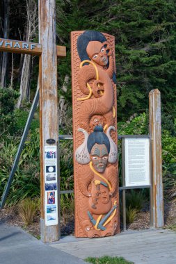 Görkemli yeşilliklerle çevrili stilize edilmiş insan yüzleri ve figürlerinden oluşan büyük, karmaşık oyulmuş ahşap bir totem direği. Direk bir ağaç zeminine ve bir patikaya dayanıyor ve yakınlarda bilgilendirme işaretleri var..
