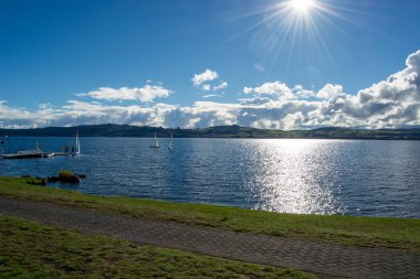 Parlak güneşin altında süzülen yelkenli tekneleriyle sakin bir göl manzarası. Gökyüzü parçalı bulutlu, sakin bir atmosferi yansıtıyor. Sahil boyunca uzanan bir patika manzarayı güzelleştiriyor..