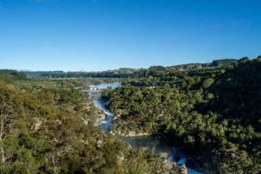 Bereketli yeşil bir manzarayla çevrili dolambaçlı bir nehir, açık mavi gökyüzünün altındaki ağaçlar ve tepeler.
