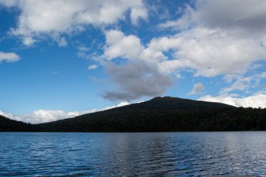 Gökyüzünü yansıtan sakin bir göl, arka planda parçalı bulutlu koşullar altında bir dağ..