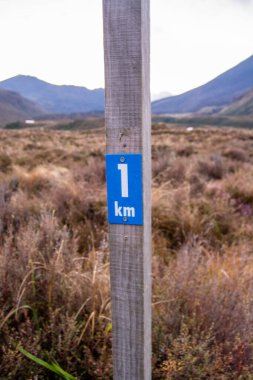 Doğal bir manzarada '1 km' yi gösteren mavi işaretli ahşap bir direk. Arka planda dağlar ve çimenlik bir alan var. Bu da yürüyüş yolu ya da açık hava yolu olduğunu gösteriyor..