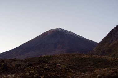 Etrafı engebeli arazi ve temiz gökyüzüyle çevrili karla kaplı görkemli bir volkanik dağ. Manzara, bölgenin doğal güzelliklerini ve jeolojik özelliklerini gözler önüne seriyor..