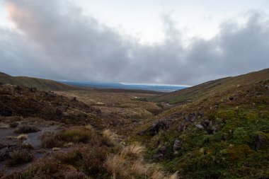 Uçsuz bucaksız bir vadinin manzarası bulutlu bir gökyüzünün altında yuvarlanan tepeler ve dağlarla çevrili. Manzara, doğal ve sakin bir atmosfer oluşturan çimen ve kayalık arazi parçalarına sahiptir..