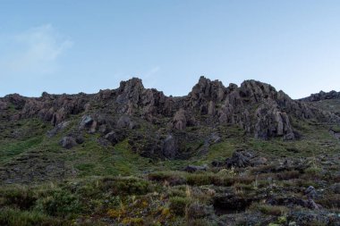 Pürüzlü kaya oluşumları içeren engebeli bir dağ manzarası ve açık mavi gökyüzünün altında yemyeşil bir bitki örtüsü..