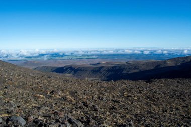Volkanik bir manzaranın panoramik görüntüsü kayalık araziyi, aşağıdaki vadiyi ve dağınık bulutlu açık mavi gökyüzünü gösteriyor..