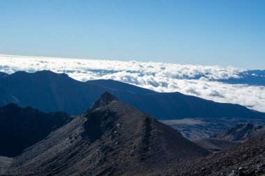 Engebeli zirveleri ve altında bulutlar olan nefes kesici bir dağ manzarası. Gökyüzü açık ve mavi, dramatik araziyi vurguluyor..