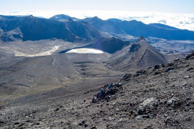 Engebeli arazinin, kraterlerin ve açık mavi gökyüzünün altındaki uzak dağların bulunduğu volkanik bir manzaranın nefes kesici görüntüsü. Sahne, doğanın saf güzelliğini yakalar ve eşsiz jeolojik oluşumları gözler önüne serer..