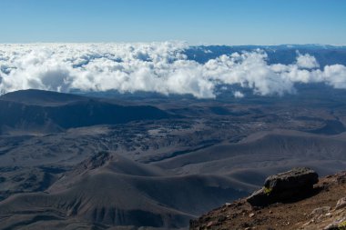 Volkanik bir zirveden nefes kesici bir manzara, engebeli arazinin engebeli bir manzarasını, yuvarlanan tepeleri ve aşağıdaki bulut denizini gösteriyor. Gökyüzü birkaç dağınık bulutla temiz, dramatik manzarayı güçlendiriyor..