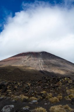 Bulutlarla kaplanmış görkemli bir volkan, engebeli kaya manzarası ve ön planda seyrek bitki örtüsü. Dağ, parlak mavi gökyüzünün altında, zirvede toprak ve kar karışımı bir görüntü sergiliyor..