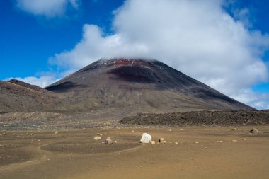 Mavi gökyüzünün altında dağınık bulutlu görkemli bir volkanik dağ. Ön planda kayalar ve kumlu bir araziyle çorak bir manzara var. Bölgenin doğal güzelliğini gözler önüne seriyor..