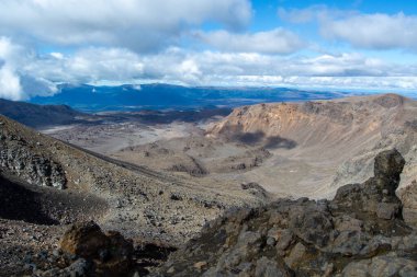 Volkanik bir manzaranın panoramik görüntüsü kayalık arazi, uzak dağlar ve bulutlu bir gökyüzü içerir. Ön planda engebeli kayalar var, arka planda ise dağınık bulutlu mavi gökyüzünün altında geniş bir vadi ve tepeler var..