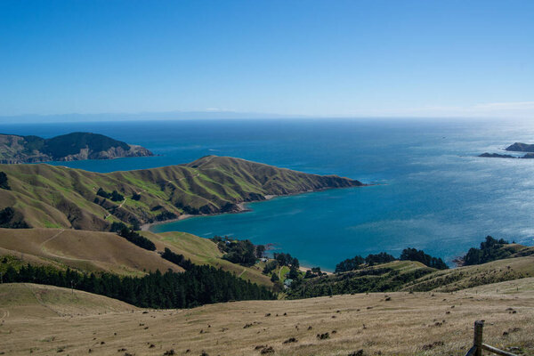 A panoramic view of a coastal landscape featuring rolling green hills, a tranquil blue sea, and distant mountains under a clear blue sky.