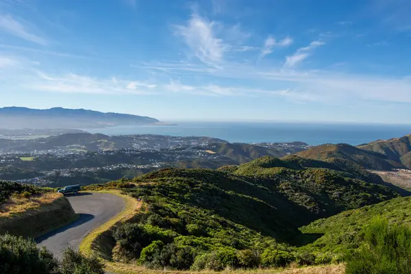 Sahil kasabası ve okyanusa bakan tepeden manzaralı bir manzara. Önünde yeşil tepeler ve açık mavi bir gökyüzü var..