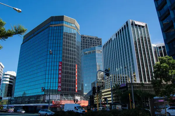Yansıtıcı cam cepheli birkaç yüksek katlı ofis binasının yer aldığı modern bir şehir manzarası. Sahne, önplanda açık mavi bir gökyüzü ve yeşil bir şehir atmosferi yaratıyor..