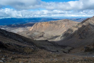 Bulutlu bir gökyüzünün altında dramatik uçurumlar ve vadilerle engebeli bir dağ manzarası. Arazide kayalık yamaçlar ve doğanın güzelliğini gözler önüne seren uzak dağlar var..