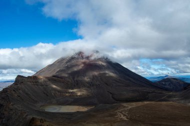 Görkemli bir volkanik dağ kısmen bulutlarla kaplı, üzerinde açık mavi bir gökyüzü var. Manzara, engebeli bir arazi ve tabanında bölgenin doğal güzelliğini gösteren küçük bir göl barındırıyor..