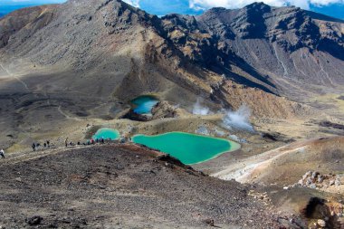 Engebeli arazi ve dağlarla çevrili canlı turkuaz göllerin yer aldığı volkanik bir manzaranın havadan görüntüsü. Yürüyüşçüler göllere giden bir patikada görülebilir, bölgenin doğal güzelliğini ve jeolojik özelliklerini sergilerler..