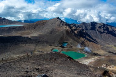 Etrafı engebeli arazi ve bulutlu bir gökyüzü altında dağlarla çevrili turkuaz göller içeren volkanik bir manzaranın nefes kesici görüntüsü..