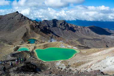 Etrafı engebeli dağlarla ve açık mavi gökyüzüyle çevrili canlı yeşil göllerin bulunduğu volkanik arazinin nefes kesici manzarası. Yürüyüşçüler ön planda görünür, manzaranın ölçeğini gösteriyor.