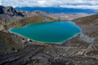 Bulutlu bir gökyüzünün altında kayalık arazi ve dağlarla çevrili çarpıcı bir turkuaz göl. Gölün canlı rengi, koyu volkanik kayalar ve arka plandaki geniş manzarayla çelişiyor..