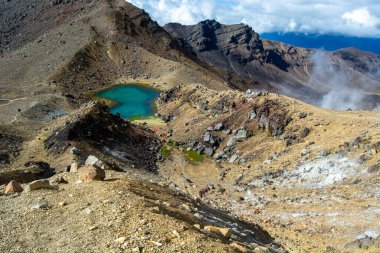 Kayalık arazi ve dağlarla çevrili turkuaz bir göl içeren volkanik bir manzaranın nefes kesen görüntüsü. Gökyüzü parçalı bulutlu, dramatik manzarayı güçlendiriyor.