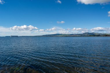 Durgun bir göl ya da deniz manzarası parlak mavi bir gökyüzünün altında, tüylü beyaz bulutlarla. Uzak tepeler sakin atmosferi artırmak için manzaralı bir zemin sağlar..