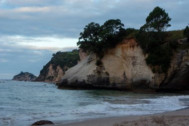 Uçurumlarda yemyeşil bir kayalık sahil manzarası, kıyıda hafif dalgalar, ve bulutlu bir gökyüzü..