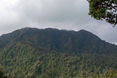 Bulutlu bir gökyüzünün altındaki yemyeşil dağ sırasının manzarası. Dağlar yoğun bitki örtüsüyle kaplı, doğanın güzelliğini gözler önüne seriyor..