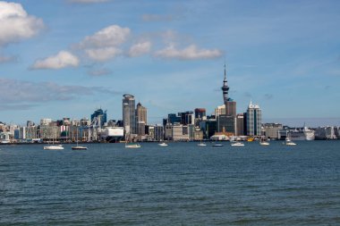 Su kenarındaki modern şehir siluetinin panoramik görüntüsü, yüksek binalar ve belirgin bir kule içeriyor. Küçük tekneler, birkaç bulutla berrak mavi gökyüzünün altında denize dağılmış durumda..