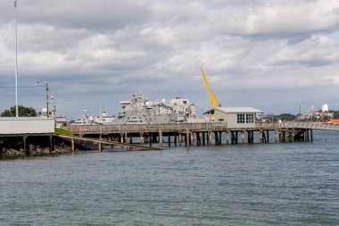 Suya uzanan bir rıhtımın manzarası. Yakınlarda bir askeri gemi var. Gökyüzü parçalı bulutlu ve arka planda bir vinç görünüyor. Bu da sahnenin endüstriyel hissini arttırıyor..