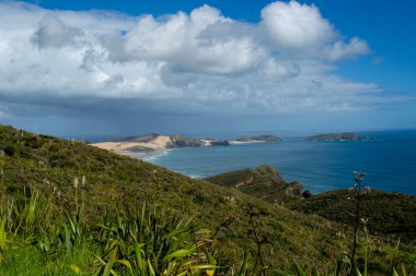 Bereketli yeşil tepeler, kumlu plajlar ve parçalı bulutlu bir gökyüzünün altında canlı mavi bir okyanus içeren manzaralı bir kıyı manzarası. Manzara bitki örtüsü ve engebeli arazinin karışımını sergiliyor..