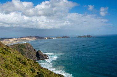 Parçalı bulutlu bir gökyüzünün altında engebeli uçurumlar, kumlu plajlar ve uçsuz bucaksız bir okyanus içeren manzaralı bir sahil manzarası. Manzara, kıyı şeridinin doğal güzelliğini nazik dalgalarla gözler önüne seriyor..