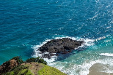 Turkuaz okyanus sularıyla çevrili kayalık bir çıkıntının manzarası. Kıyıda hafif dalgalar çakıyor. Kıyı şeridinde yemyeşil bitki örtüsü ve kumlu plaj alanları yer alıyor..